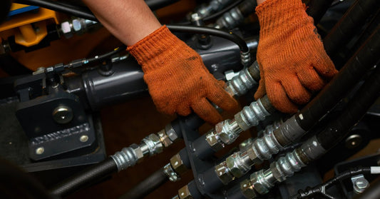 Orange-gloved hands adjusting black hydraulic hoses and metal fittings inside a truck service compartment.
