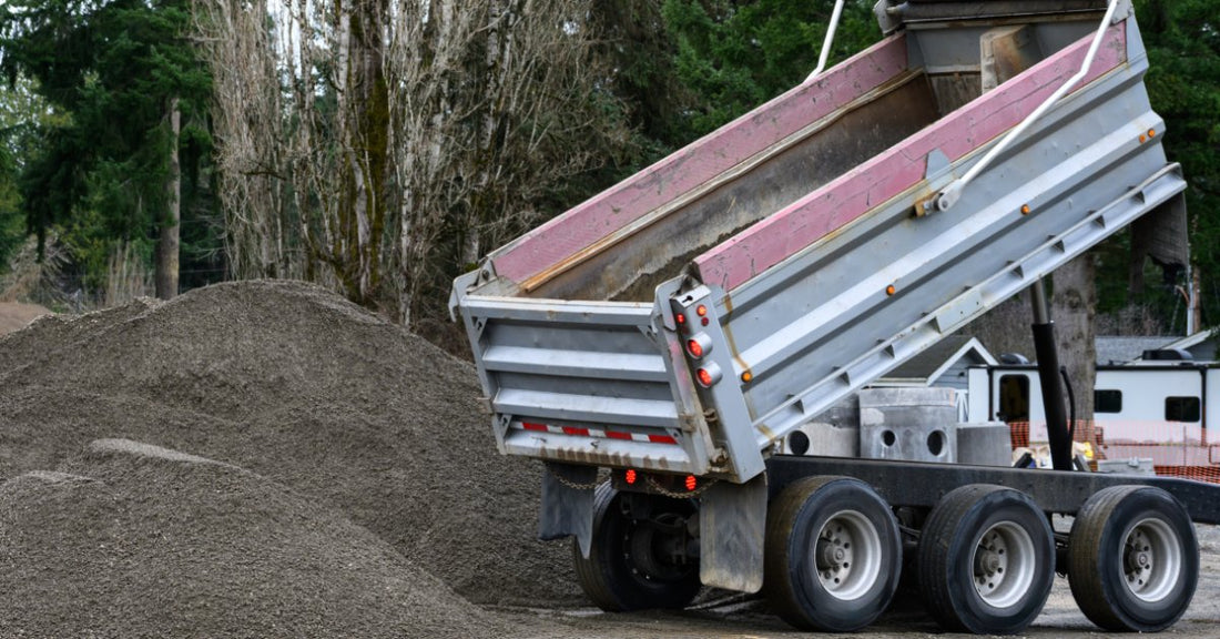 A dump truck with a raised bed unloading gravel onto a large pile near trees, trailers, and work barriers.