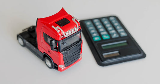 A toy cargo truck sitting beside a calculator, paper bills, and coins on a tabletop with paperwork nearby.