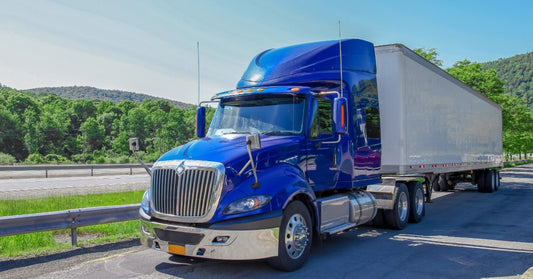 A blue semi truck with a white trailer parked beside a highway, with trees and hills under a clear sky.