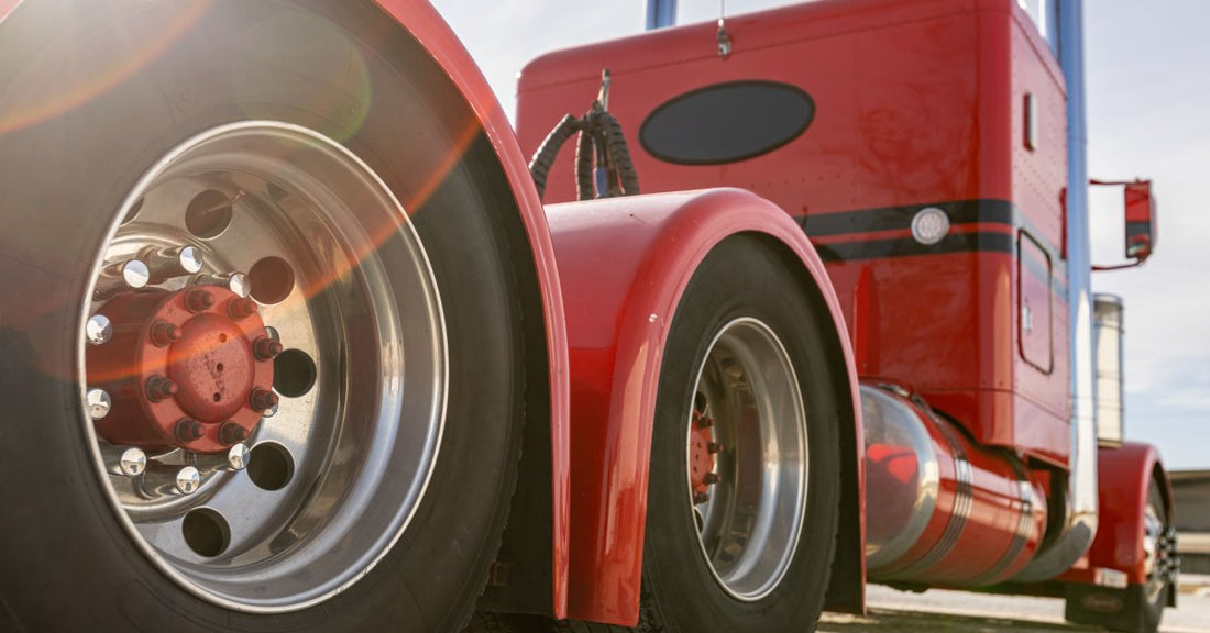 A dynamic, low-to-the-ground shot of a classic red semi-truck with matching red axles, and a lens flare effect in the corner.