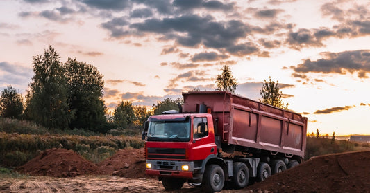 A red dump truck with a matching bed rolling through a small field of dirt, past a few mounds of dirt.