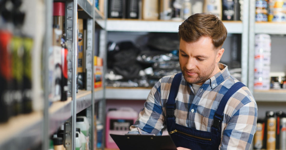 A man in work overalls writing on a clipboard between stocked shelves of automotive bottles and supplies.