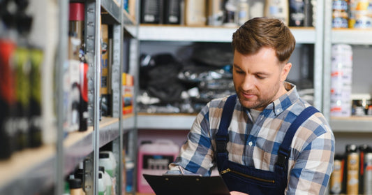 A man in work overalls writing on a clipboard between stocked shelves of automotive bottles and supplies.