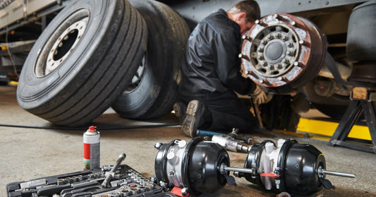 A man in a black leather jumpsuit working on the wheels of a large semi-truck, surrounded by various tools and spare parts.