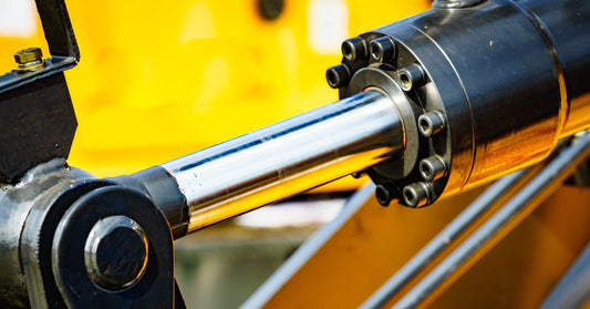 A close-up of a polished steel hydraulic cylinder in a black case, mounted on a bright yellow machine.