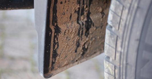 A close-up of a one of a silver vehicle's mud flaps, which is almost entirely caked in thick, brown mud.