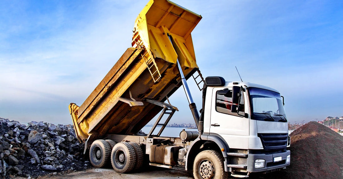 A yellow dump truck with its bed tilted backward, unloading rocks onto the ground beneath a clear blue sky.