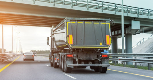 The back view of a truck with a dump trailer attached to it as it drives underneath a bridge over the road.