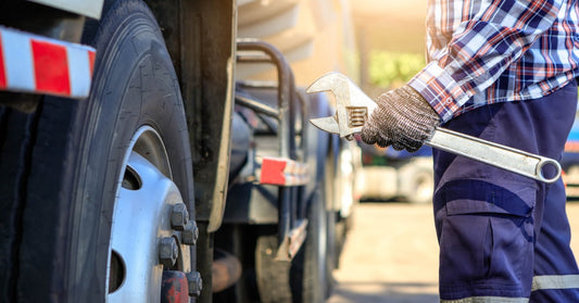 A man wearing a plaid shirt, blue pants, and gloves holding a large wrench and standing besides a semi-truck's front wheels.