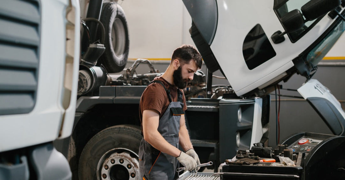  A man wearing overalls standing at a workbench underneath a propped-up semi-truck with it's hood open.