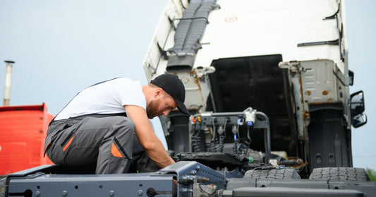 A mechanic kneeling on the bed of a lifted truck trailer while repairing part of the vehicle outdoors.