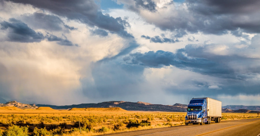 A semi-trailer truck driving along a highway road with a colorful sunset sky and clouds in the background.