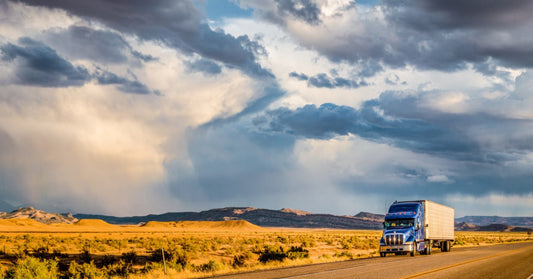 A semi-trailer truck driving along a highway road with a colorful sunset sky and clouds in the background.