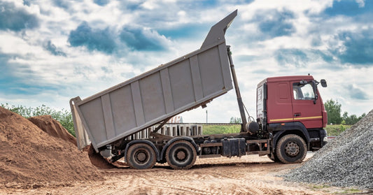 A dump truck with its raised bed unloading material at a construction site with dirt and equipment visible nearby.
