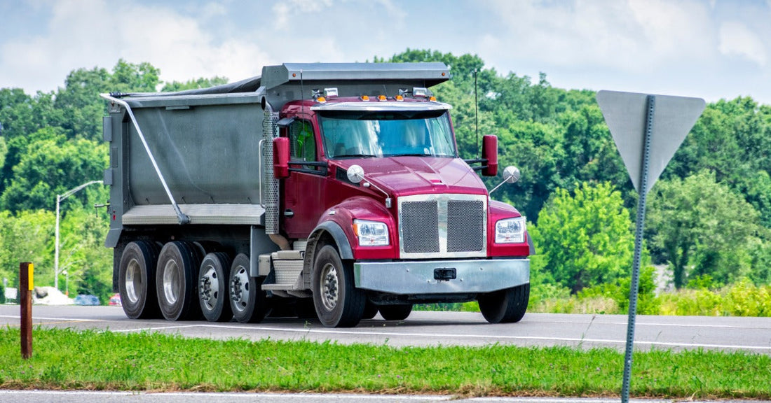 A red dump truck driving along a paved road with a forest visible behind the vehicle in the background.