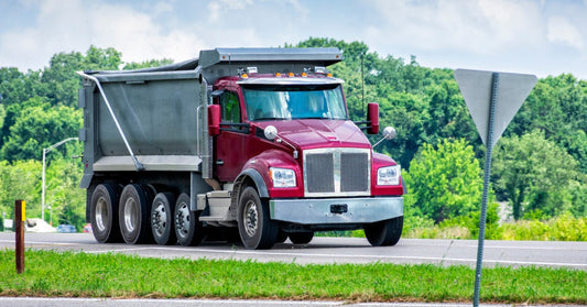A red dump truck driving along a paved road with a forest visible behind the vehicle in the background.