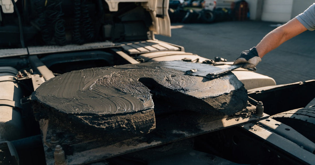 A worker applying lubricant to a semi-truck fifth wheel coupling during mechanical maintenance outdoors.