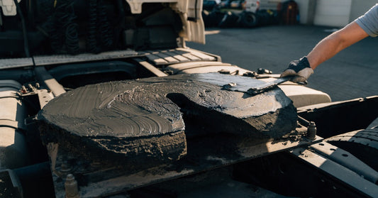A worker applying lubricant to a semi-truck fifth wheel coupling during mechanical maintenance outdoors.