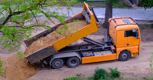A large orange dump truck unloading sand onto a dirt area near industrial equipment under a clear blue sky.