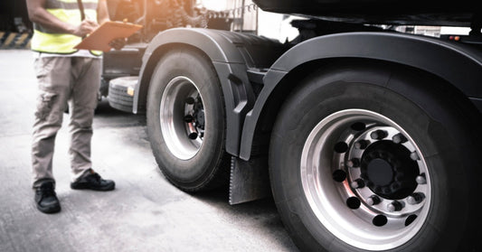 A person standing outside beside a semi-truck tire while holding a clipboard, with the sun shining down from above.