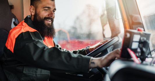 A bearded worker in an orange safety jacket sitting inside a truck cab, smiling while holding the steering wheel.