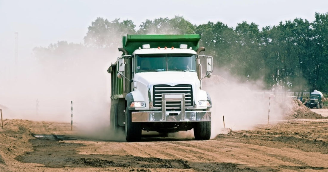 A white and green dump truck driving on a dusty dirt road with trees and construction materials in the background.