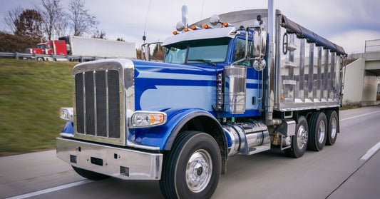 A blue and silver dump truck driving on a highway lane, with a red semi truck blurred in the background.