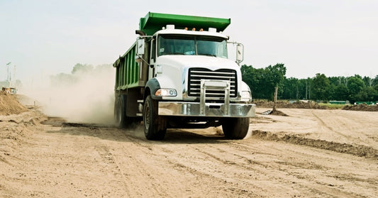 A green and white dump truck driving along a dusty dirt road with trees and open construction land behind it.