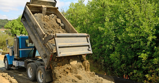 A blue dump truck unloading dirt and large rocks on a roadside area bordered by dense green trees and sloped ground.