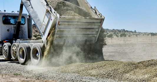 A raised dump truck bed pouring gravel onto a dusty unpaved road beside an open, dry rural landscape.