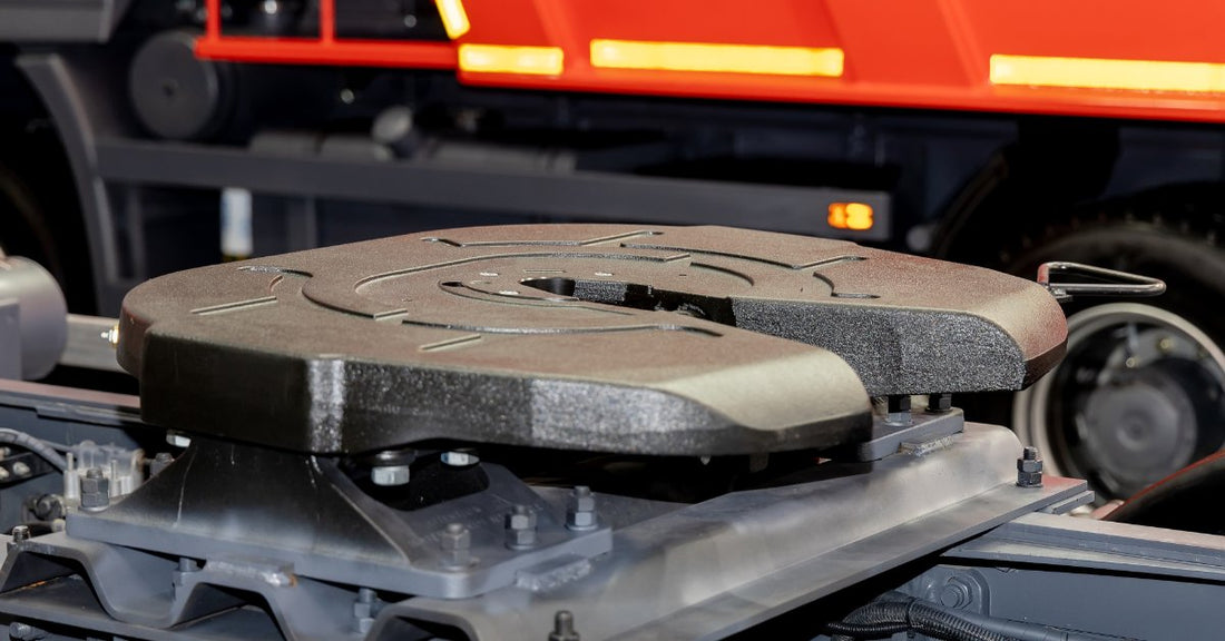 A close-up fifth wheel coupling mounted on a gray truck chassis with bolts, rails, and a red trailer behind it.