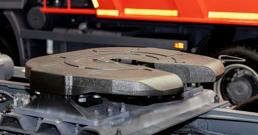 A close-up fifth wheel coupling mounted on a gray truck chassis with bolts, rails, and a red trailer behind it.